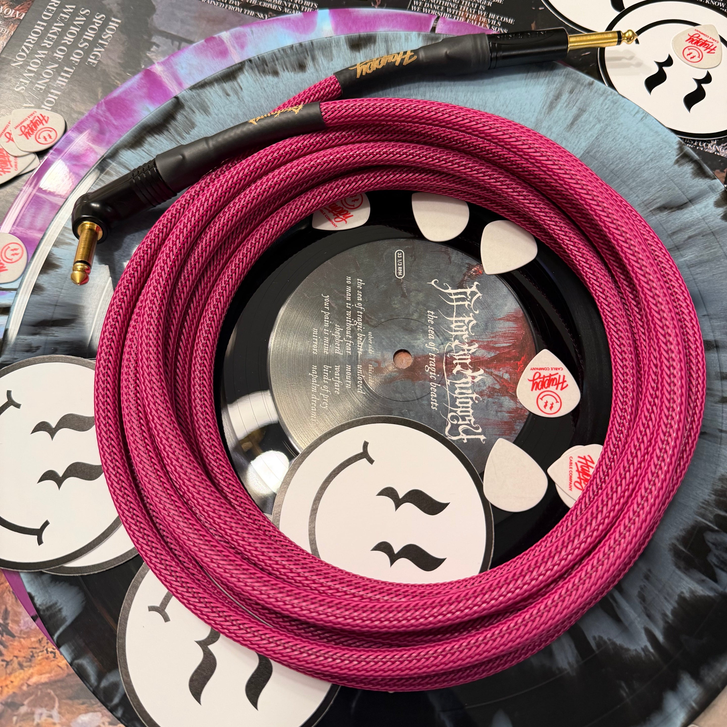 Pink low-noise guitar cable coiled on a vinyl record with a black sleeve featuring white smiley face designs.